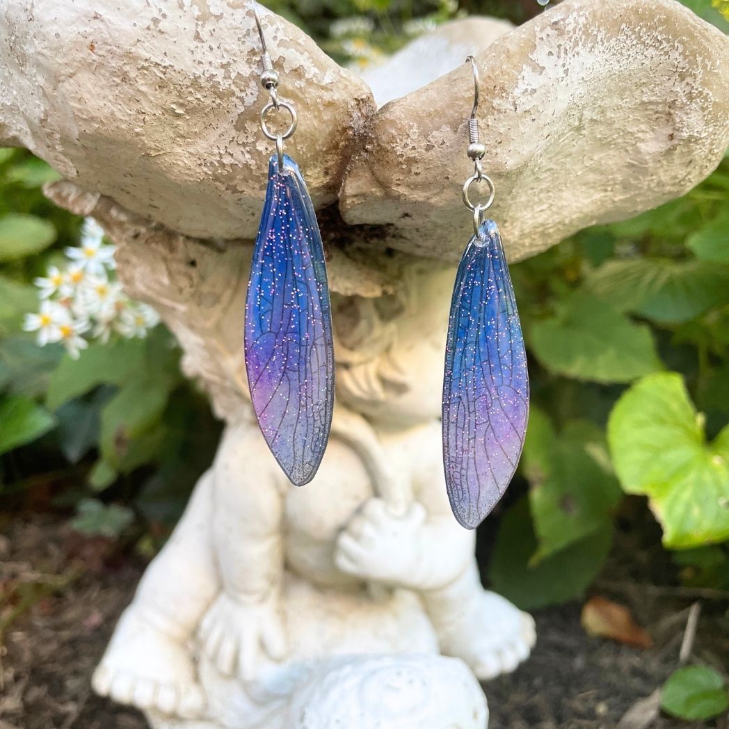 Sparkly glitter blue and purple dragonfly wing earrings hanging from a garden ornament.