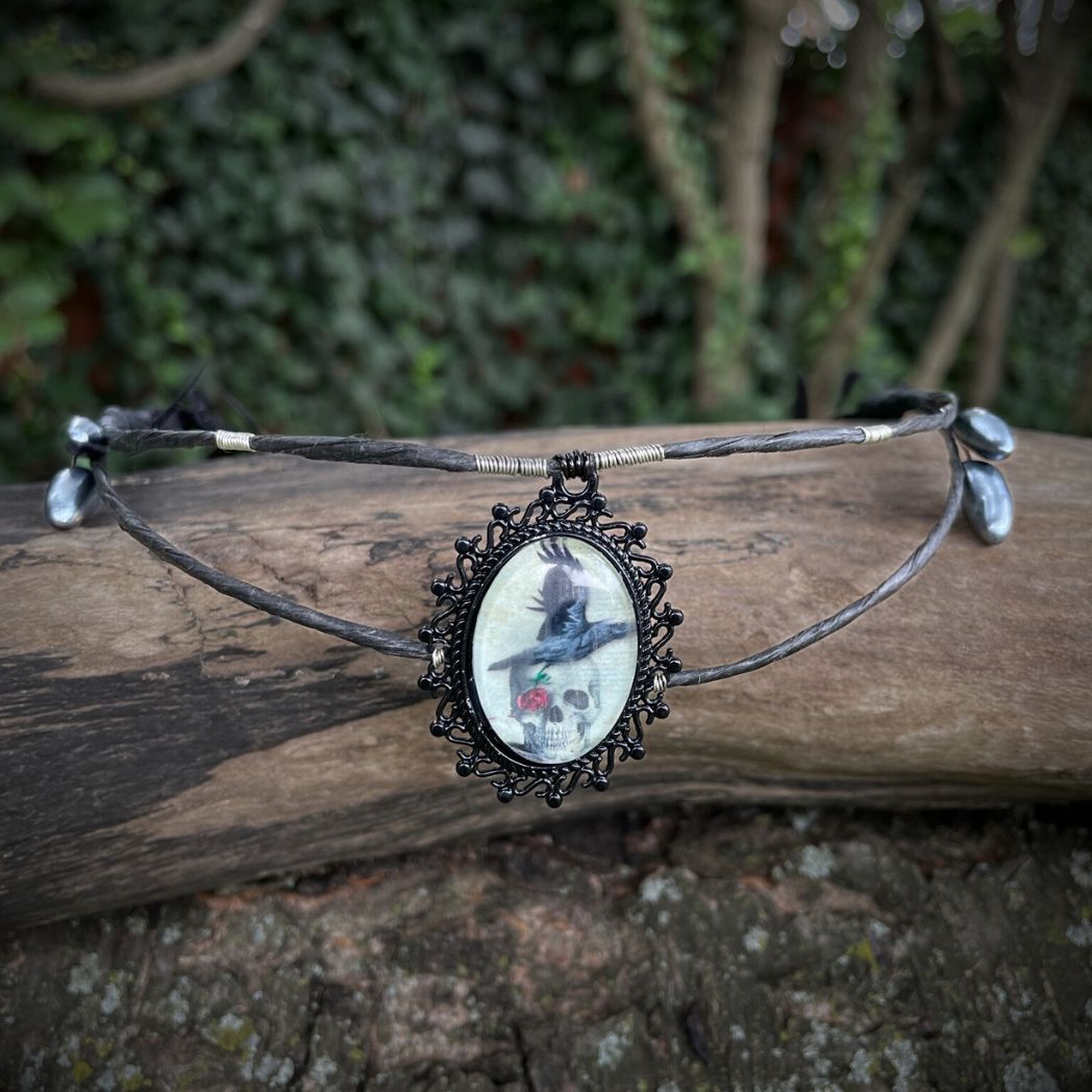 Black wire crown with oval cabochon focal of a raven and skull resting on a tree branch.