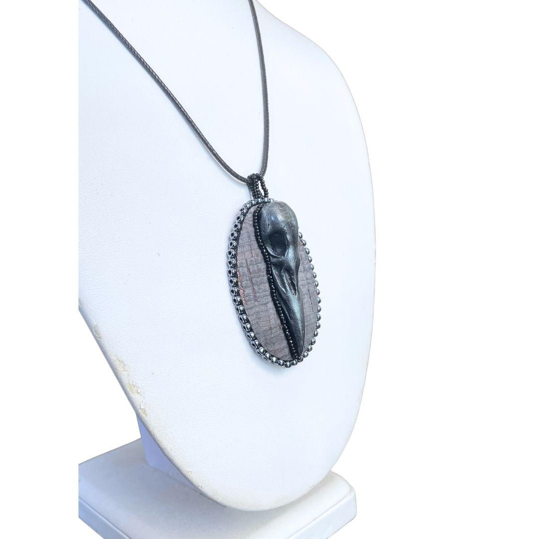 Side view of An oval pendant with a polymer clay crow skull on gray cork and hand beaded with silver and black seed beads on a display mannequin