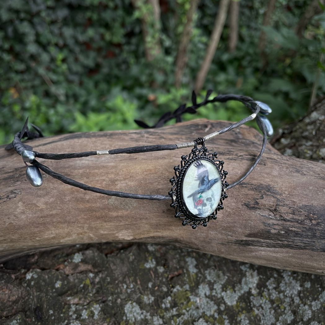 Black wire crown with oval cabochon focal of a raven and skull resting on a tree branch.