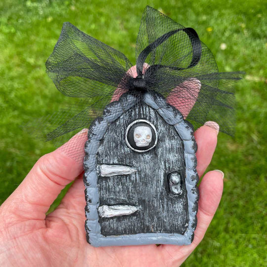 Dark fairy door ornament with skull and bones. Held in hand over grass.