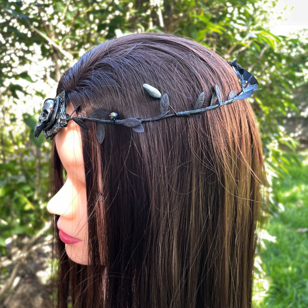 A silver raven skull on textured gold highlighted diamond shape focal on a black leaf circlet with black crystals and silver flower buds. Displayed side profile on a model 