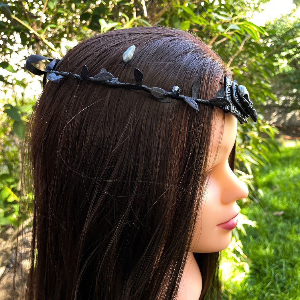 A silver raven skull on textured gold highlighted diamond shape focal on a black leaf circlet with black crystals and silver flower buds. Displayed side profile on a model
