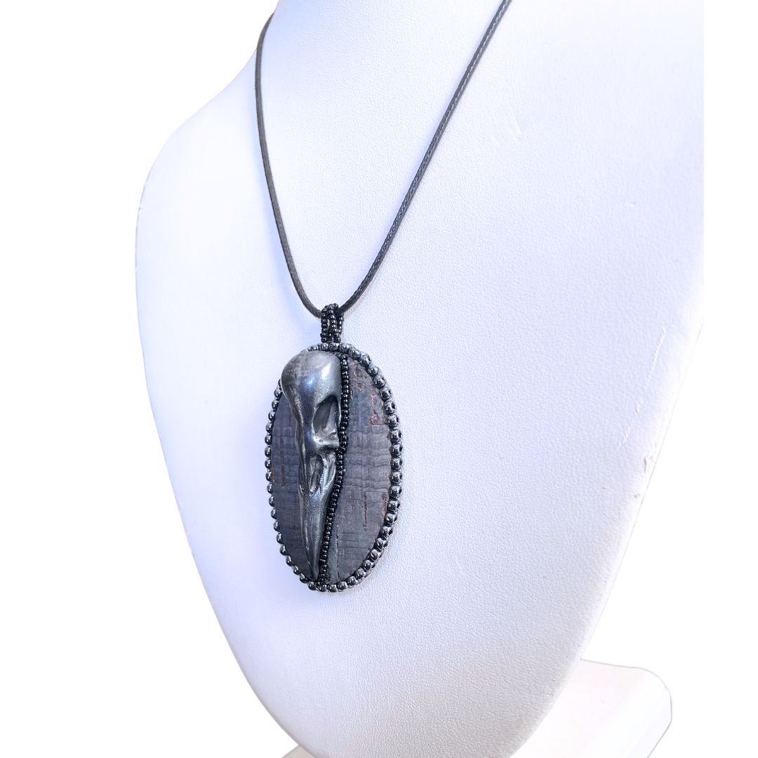 Side view of An oval pendant with a polymer clay crow skull on gray cork and hand beaded with silver and black seed beads on a display mannequin