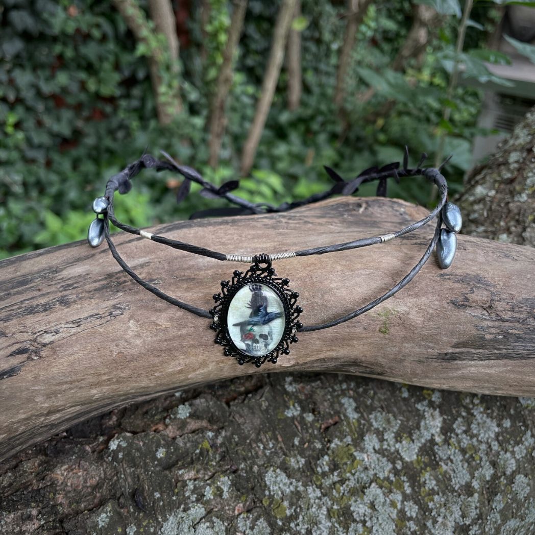 Black wire crown with oval cabochon focal of a raven and skull resting on a tree branch.