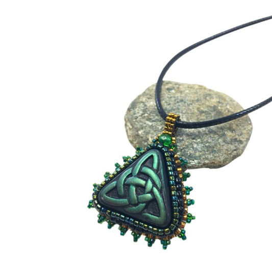 Side profile of a green trinity beaded pendant necklace resting on a pebble against a white background.