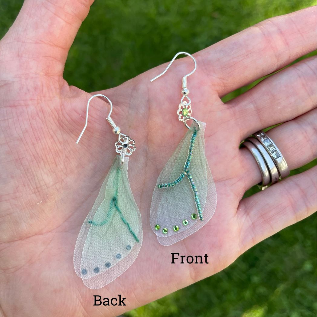 Green sheer fabric beaded butterfly wing earrings with green crystals with a flower bead connecting to the ear wires. held in a hand.