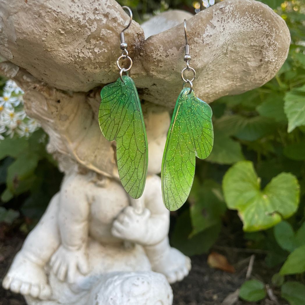 Green ombre sparkly glitter plastic fairy wing earrings hanging from a fairy garden ornament