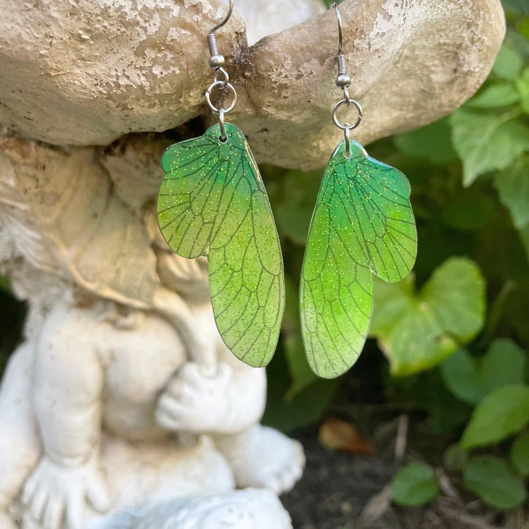 Green ombre sparkly glitter plastic fairy wing earrings hanging from a fairy garden ornament