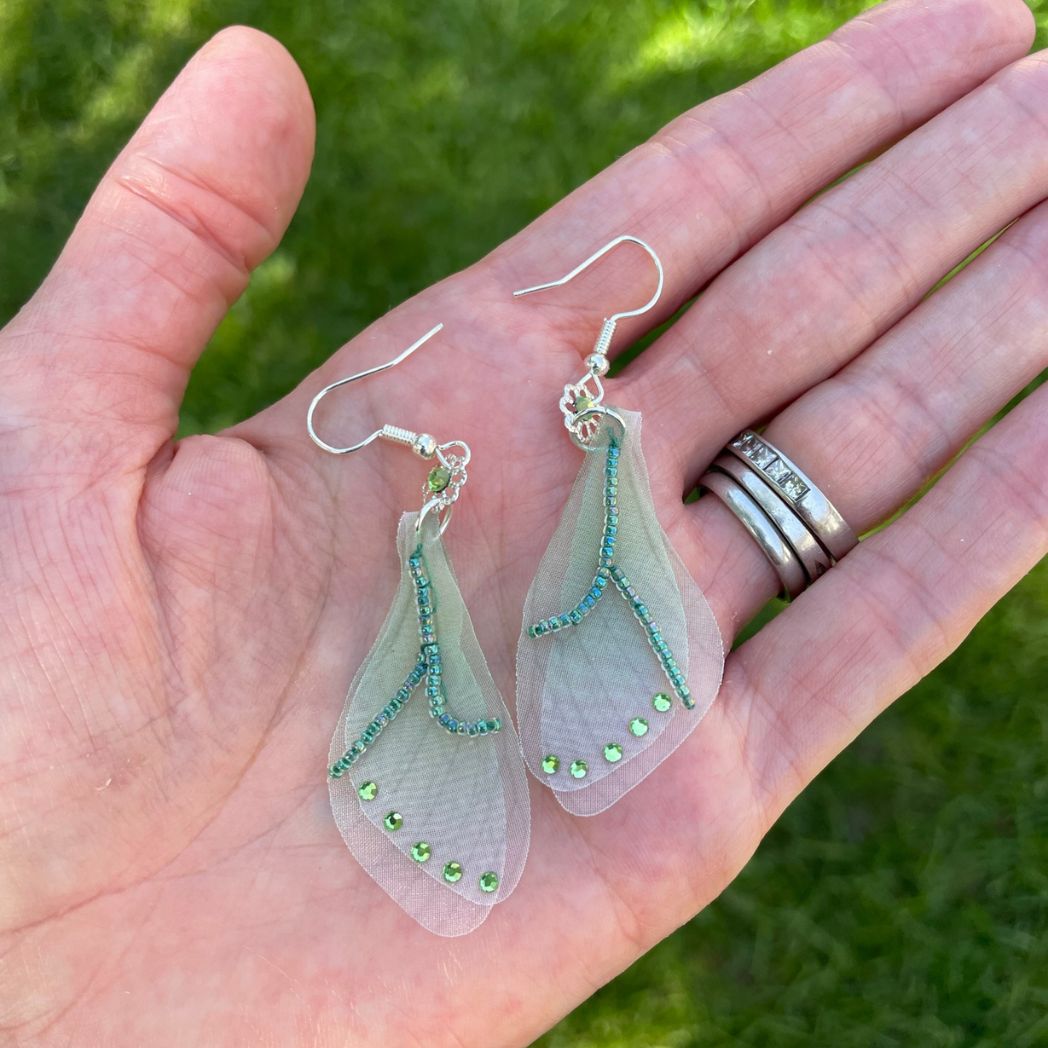 Green sheer fabric beaded butterfly wing earrings with green crystals with a flower bead connecting to the ear wires.  Held in a hand,