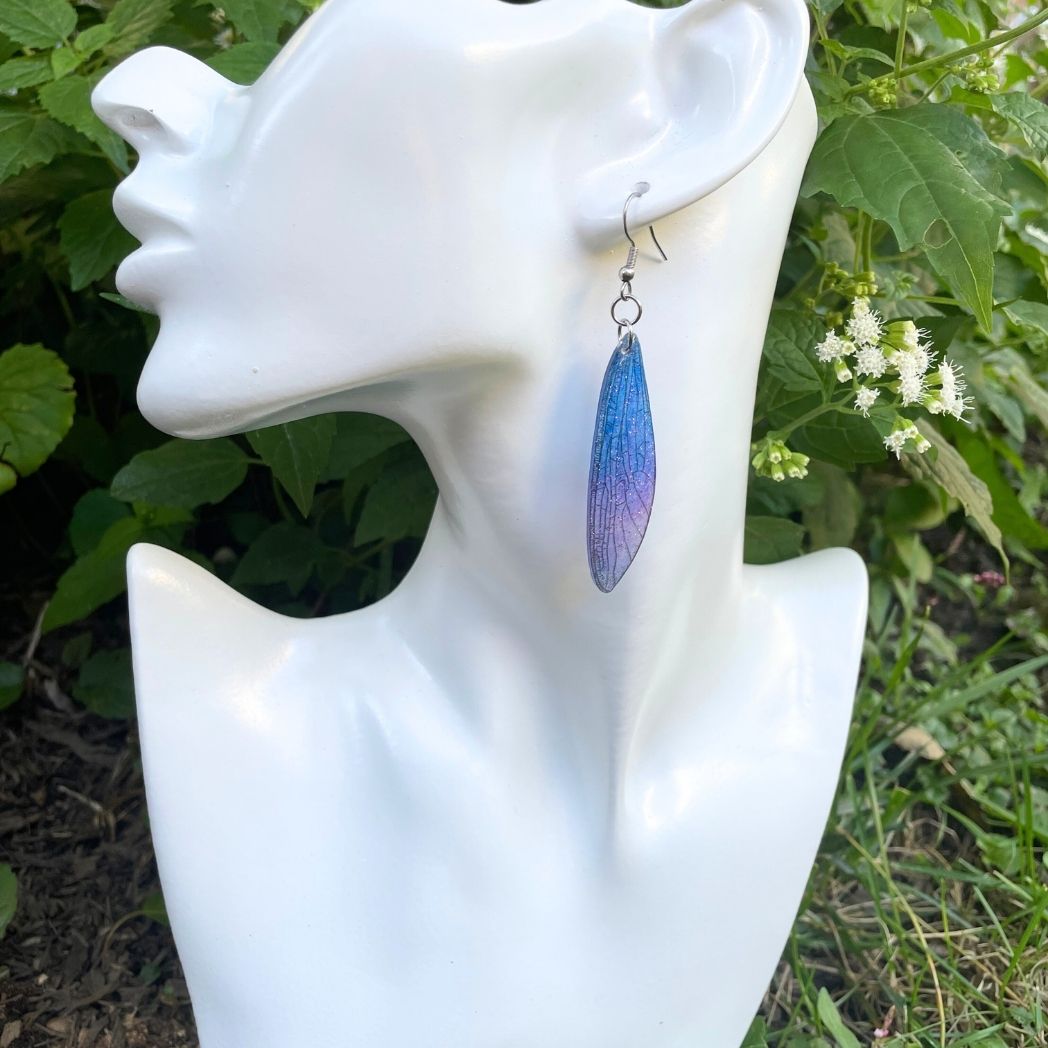 Sparkly glitter blue and purple dragonfly wing earrings hanging from an earring bust.