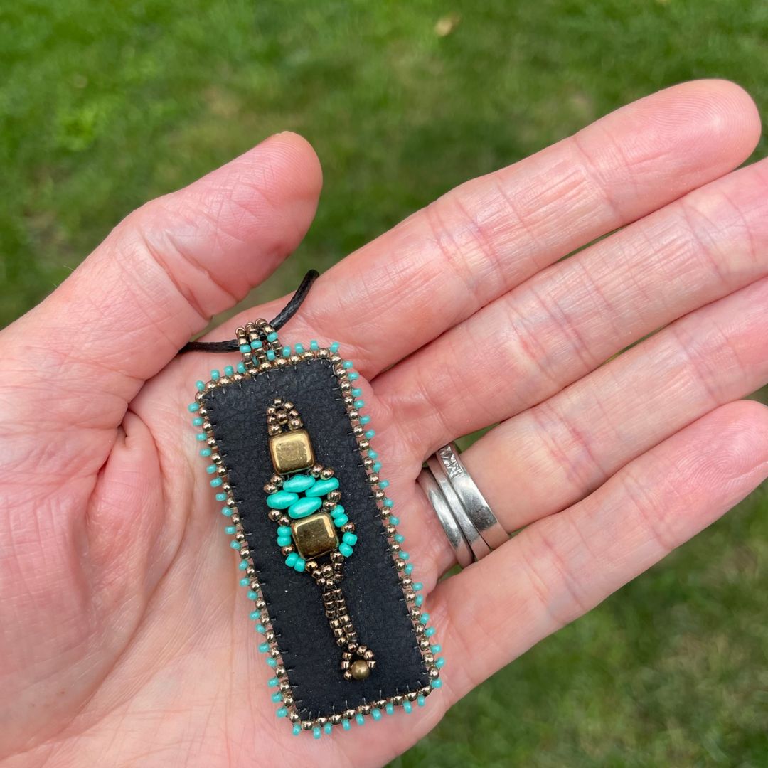 Renaissance pendant with  black faux leather rectangle pendant with turquoise and gold beading with black cord necklace. held in hand.