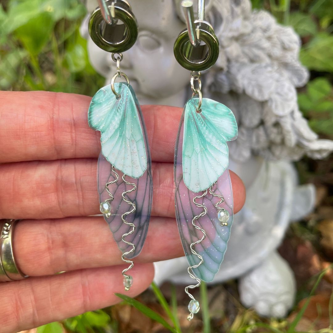 mint green butterfly jewelry earrings in a garden with a hand behind them to show the size