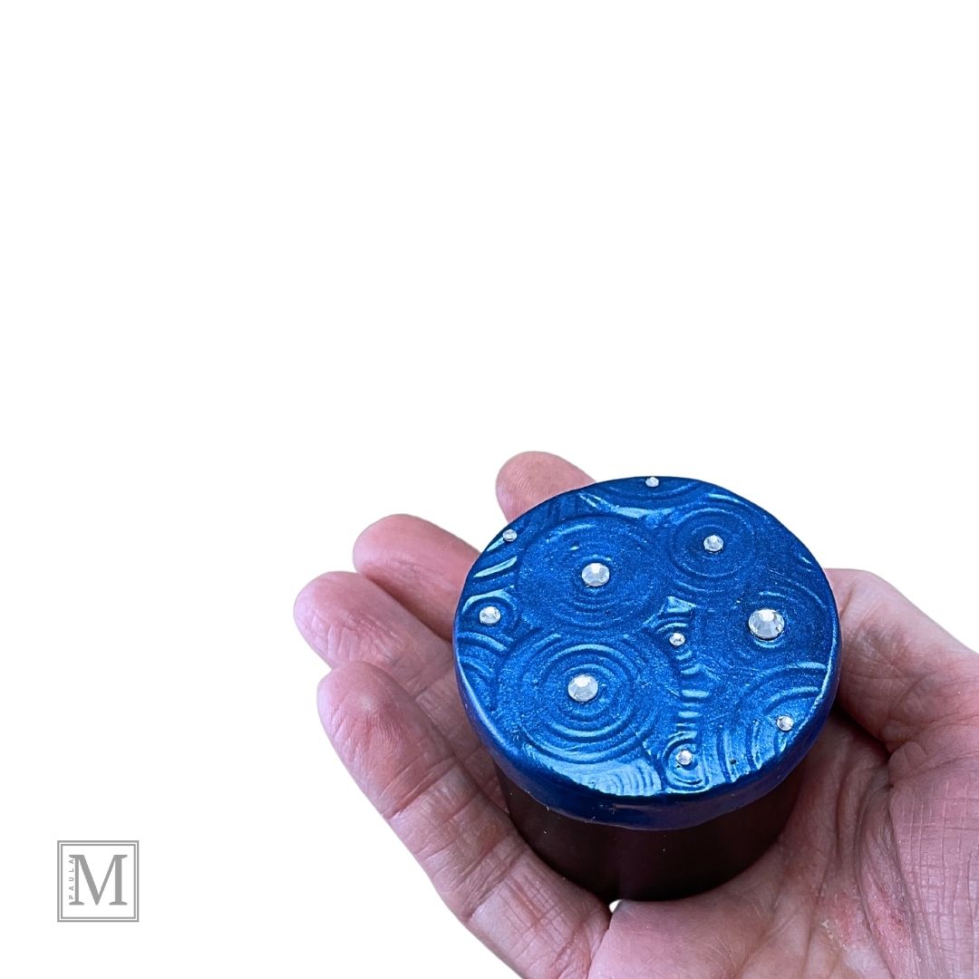 A black round tin with decorated lid. Lid is decorated with polymer clay and hand painted metallic blue with blue and clear crystals shown in hand.