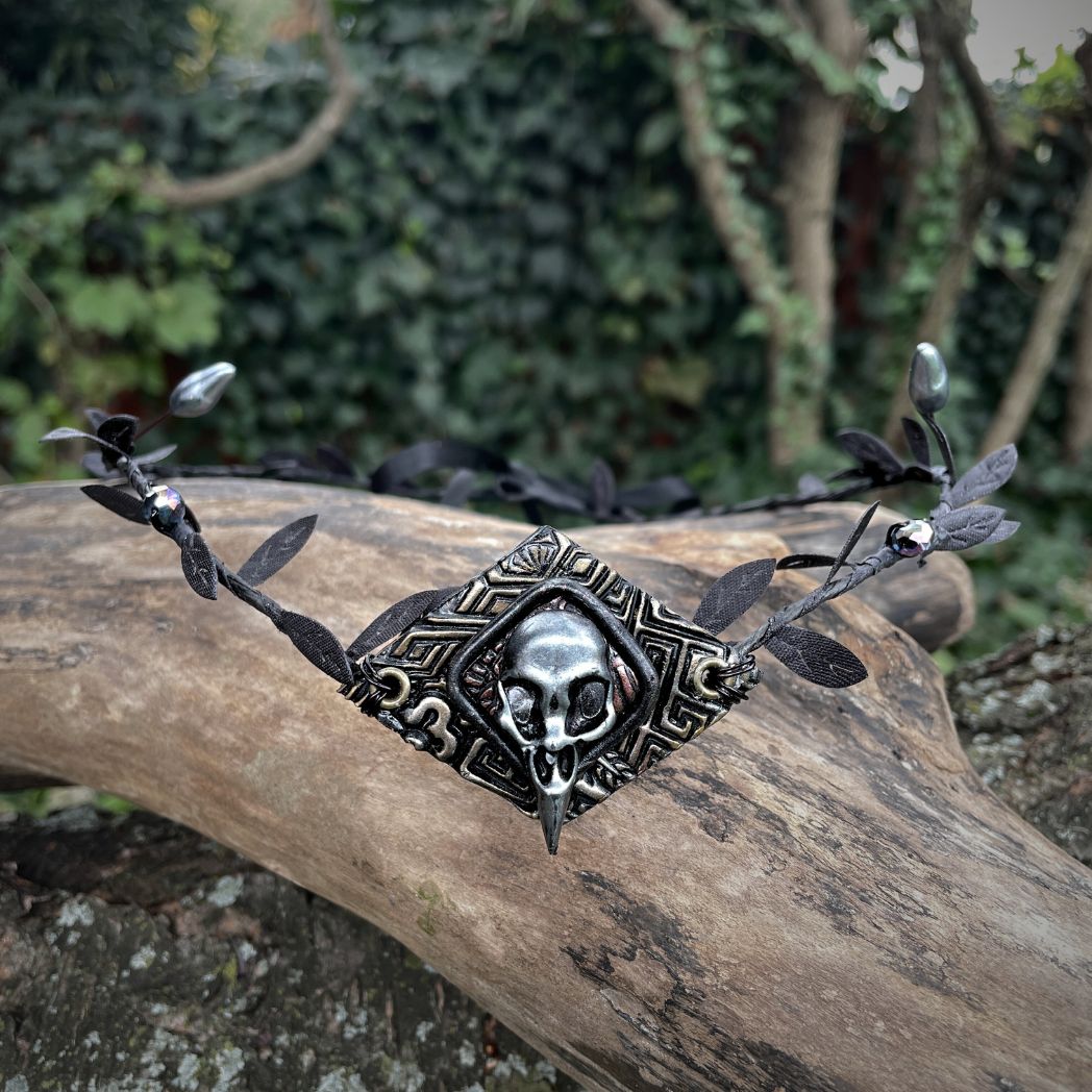 A silver raven skull on textured gold highlighted diamond shape focal on a black leaf circlet with black crystals and silver flower buds. Circlet is resting on a tree branch.