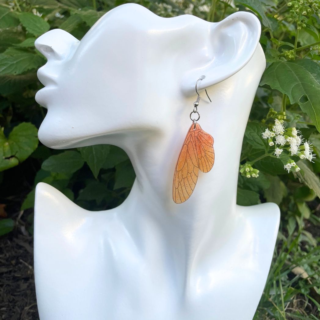 orange and yellow sparkly glitter plastic fairy wing earrings hanging from an earring bust.
