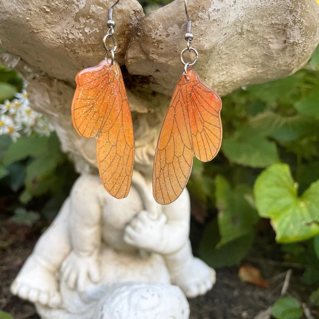 orange ombre sparkly glitter plastic fairy wing earrings hanging from a fairy garden ornament