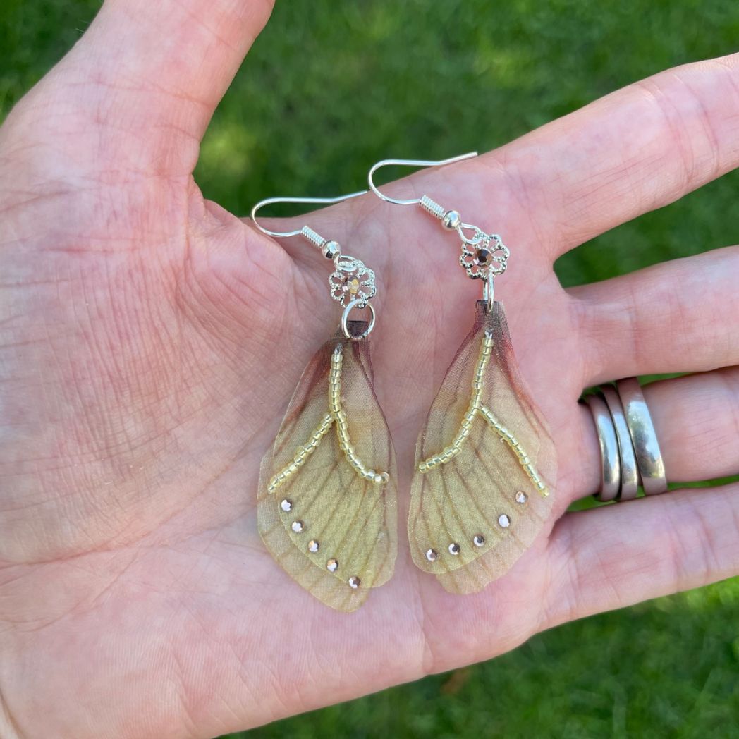 Yellow sheer fabric beaded butterfly wing earrings with peach color crystals and a flower bead connect the wings to the ear wires. Held in a hand.