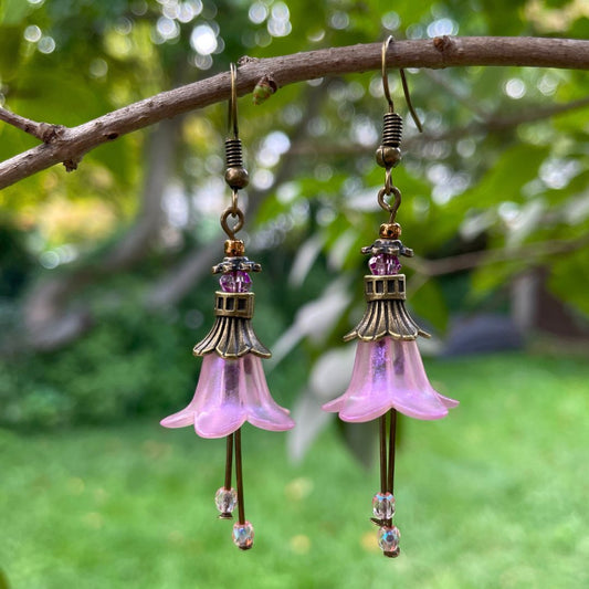 Pink Bell flower dangle  earrings with purple crystal and pink crystal stamen with antique bronze finish. Earrings are hanging from a tree branch. 