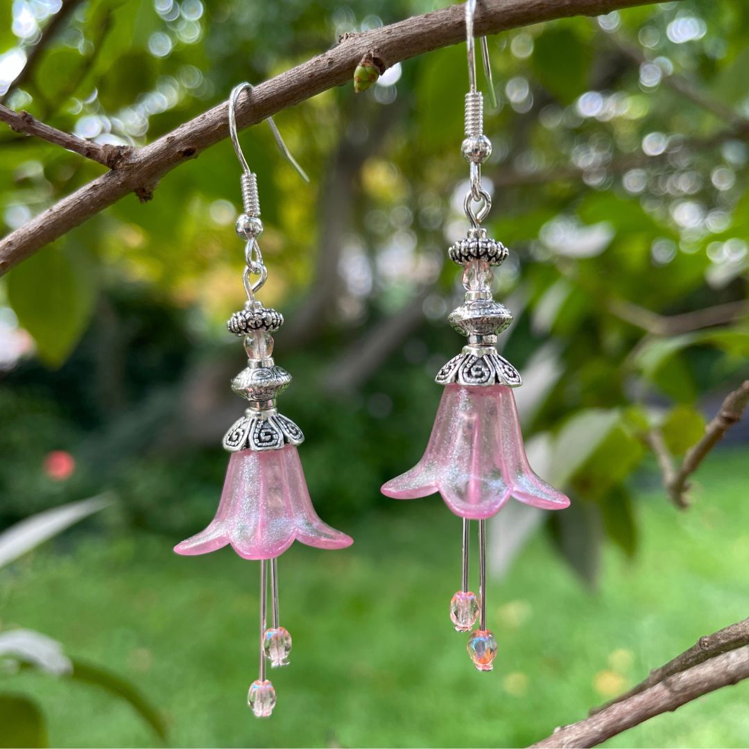 Pink Bell flower dangle  earrings with pink crystal stamen with silver finish. Earrings are hanging from a tree branch. 