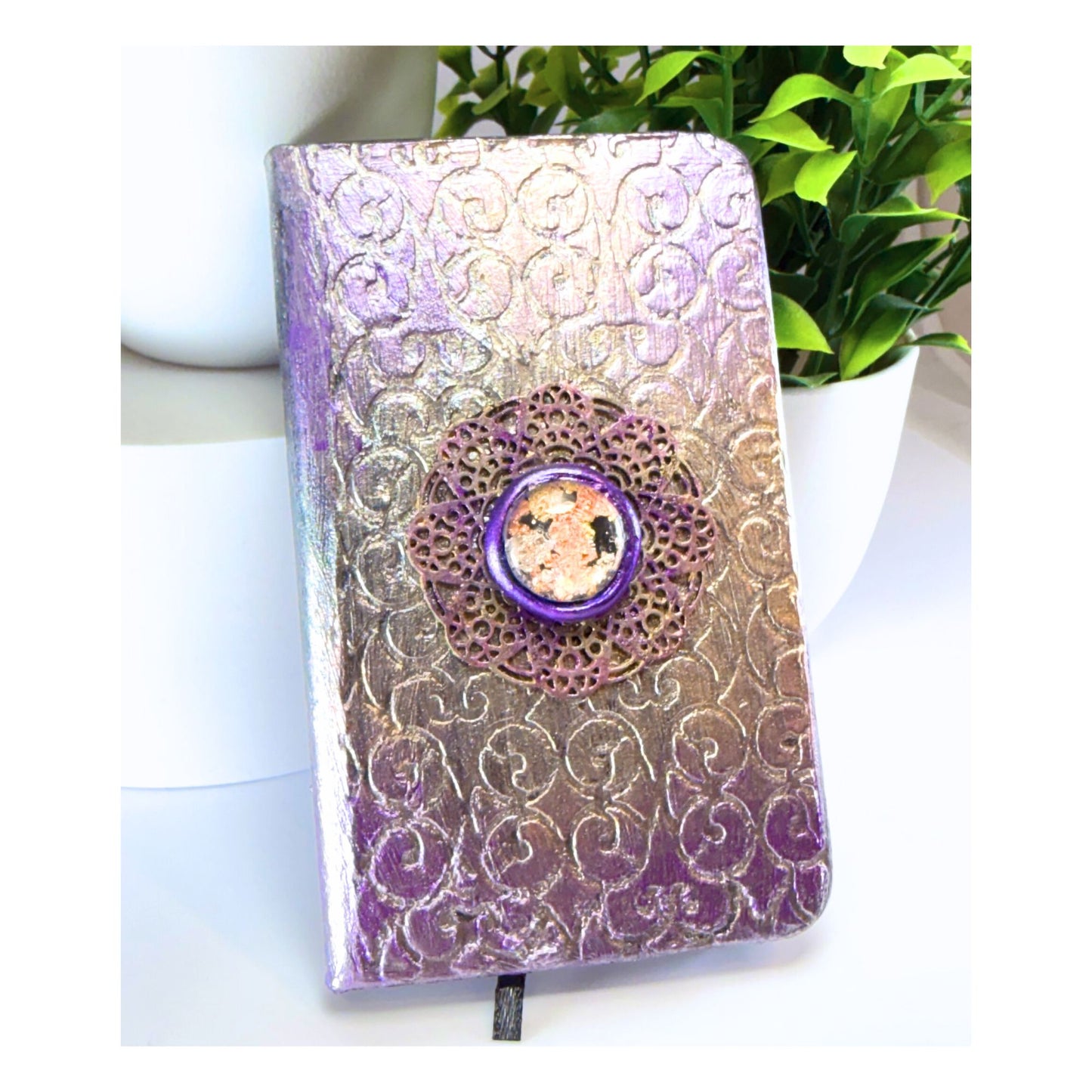 Decorative notebook with floral embossing and a purple button on a white surface with a plant in the background