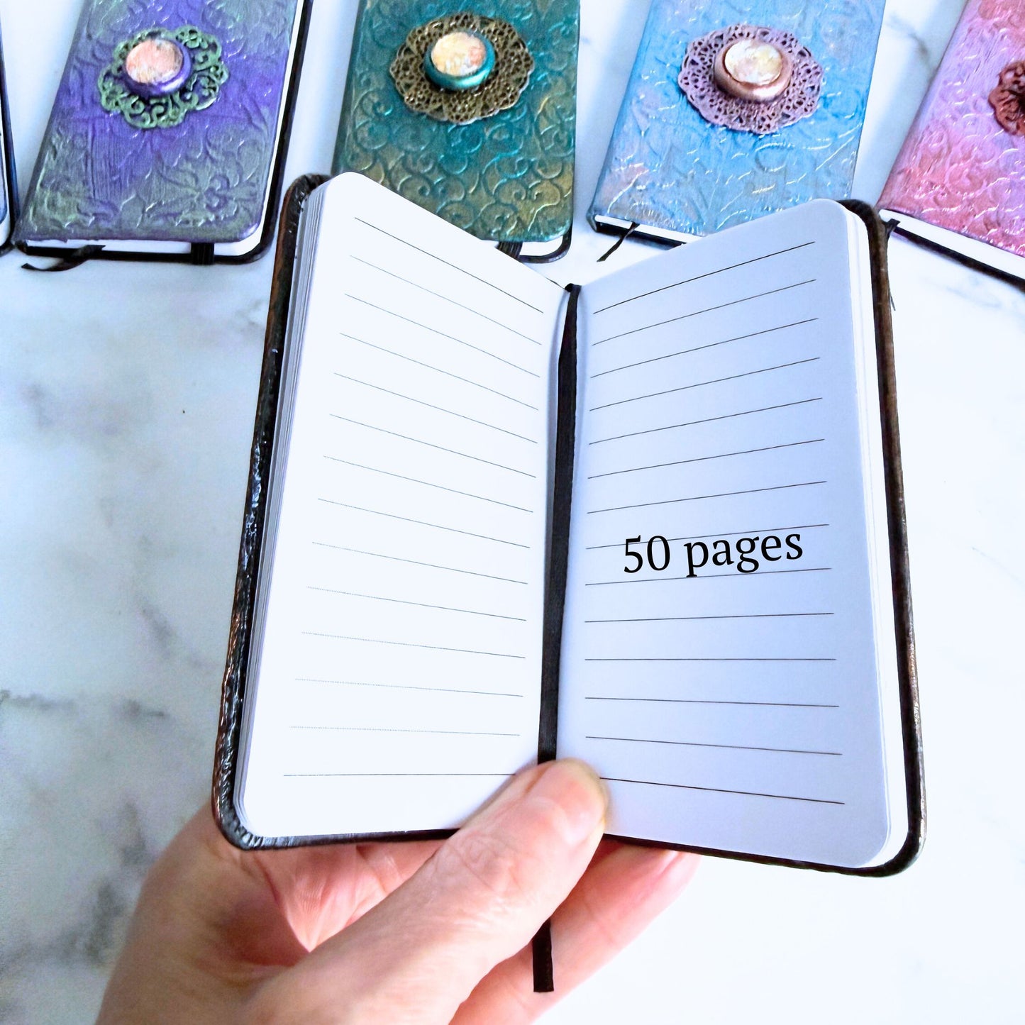 Small notebook with lined pages and a decorative cover, held open by a hand on a marble surface.