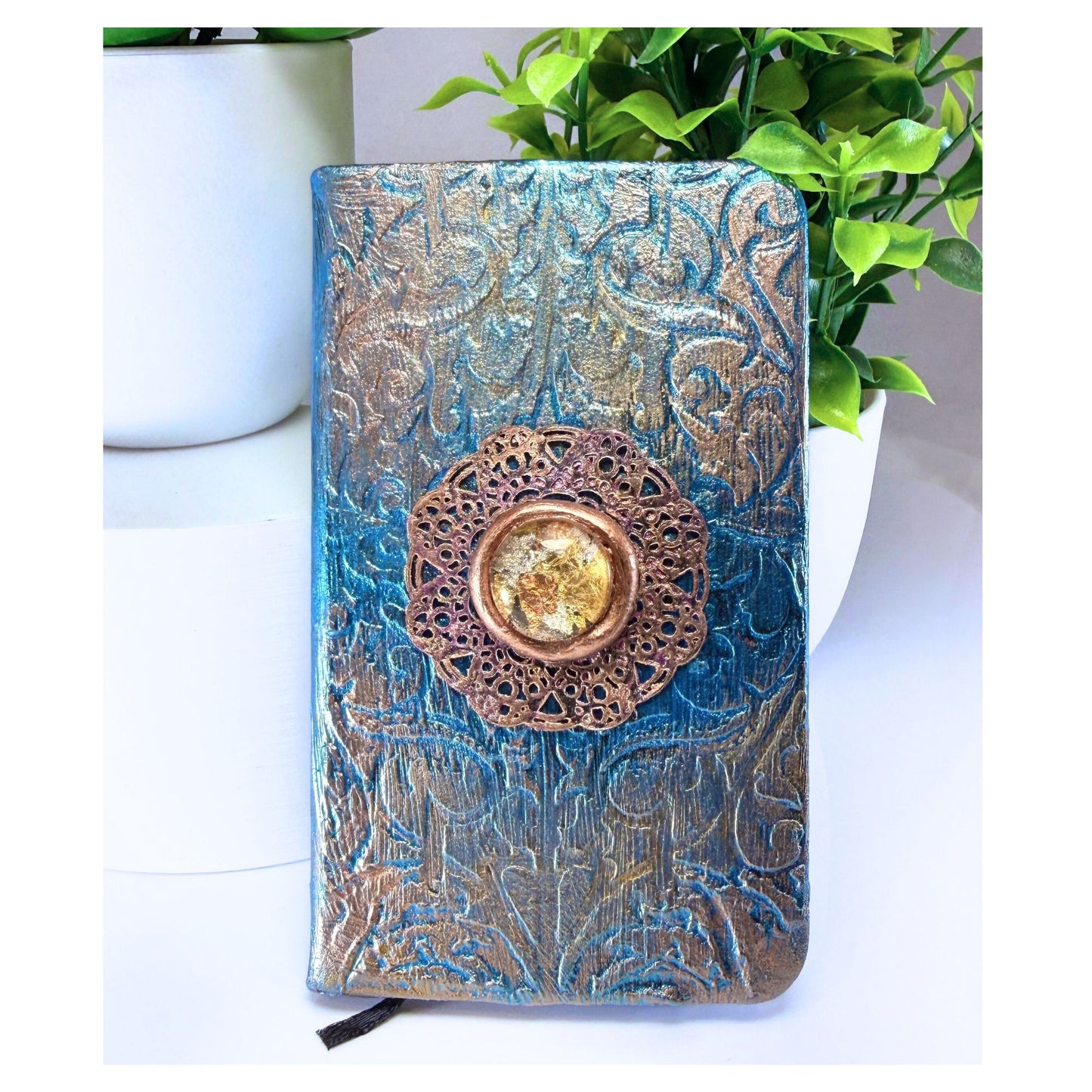 Decorative notebook with intricate design and gold embellishment on a white surface with plants in the background.