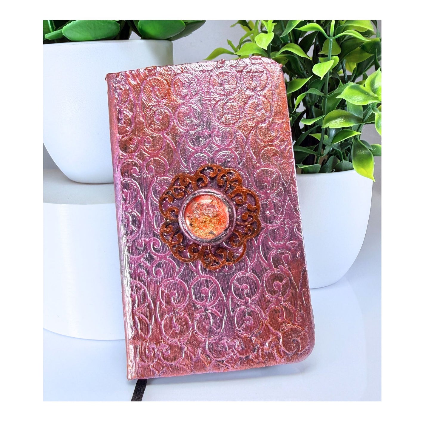 Decorative notebook with embossed design and central embellishment on a white surface with plants in the background