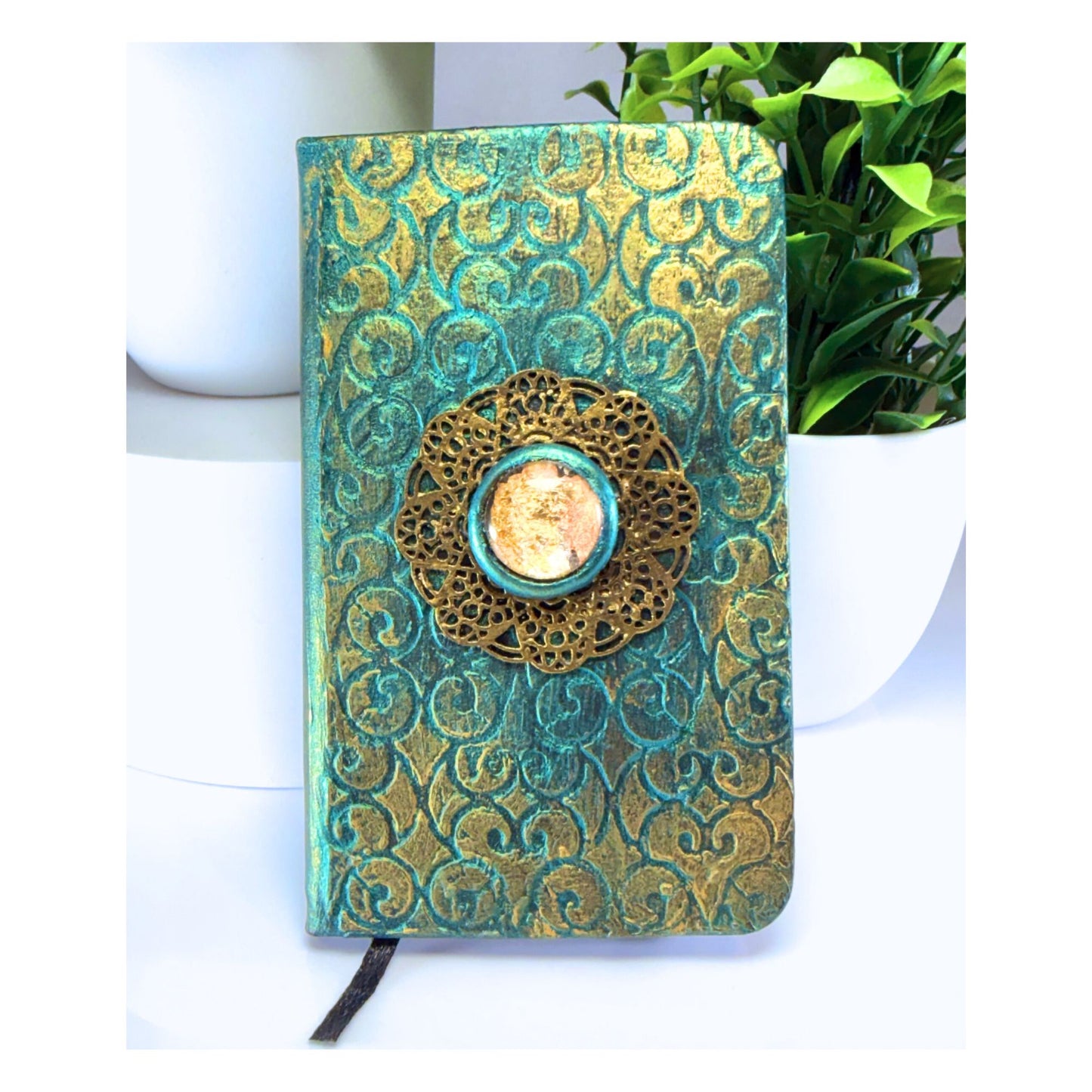 Decorative notebook with gold and teal patterns on a white surface with a plant in the background