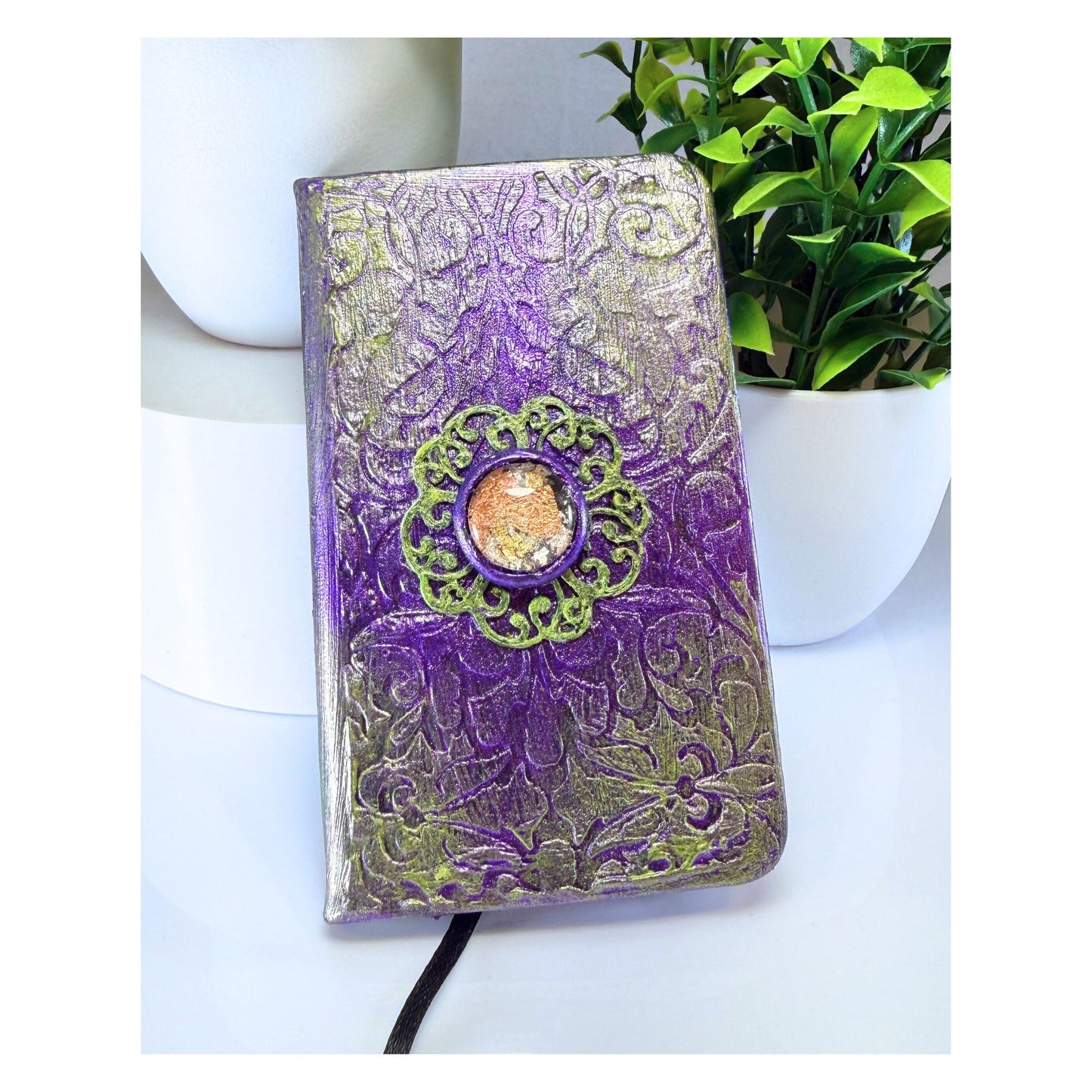 Purple and silver embossed notebook with decorative element on a white surface with a plant in the background