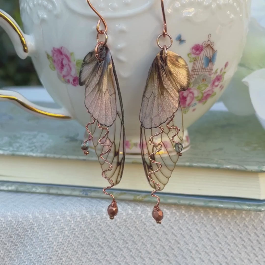 fairy grunge  brown fairy wing earrings with copper wired crystal on the side of a teacup with a hand that holds them to show the details of the the earrings.