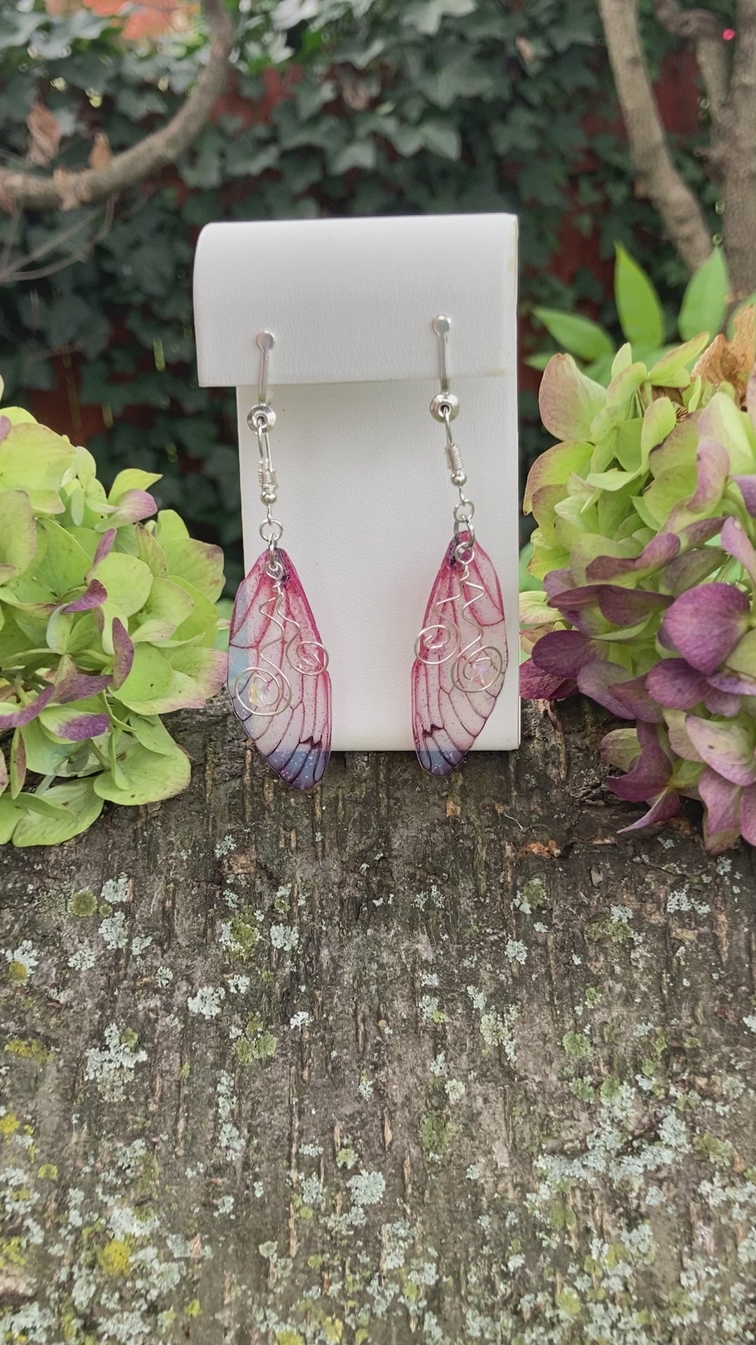 pink butterfly wing clip on earrings on an earring display outside with hydrangeas either side