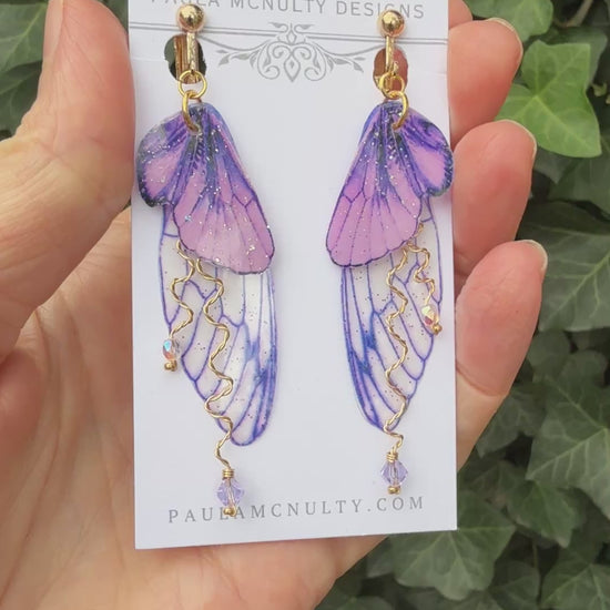 Hand holding purple clip-on butterfly wing earrings with gold finish and comfort pads by Paula McNulty Designs.