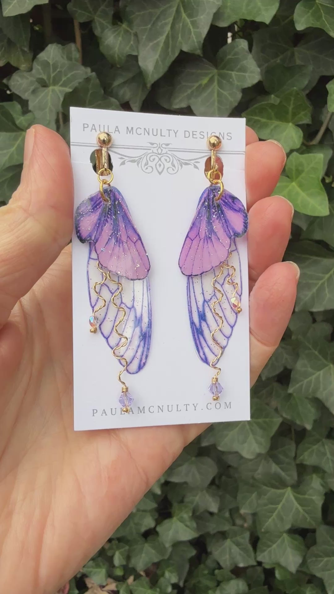 Hand holding purple clip-on butterfly wing earrings with gold finish and comfort pads by Paula McNulty Designs.