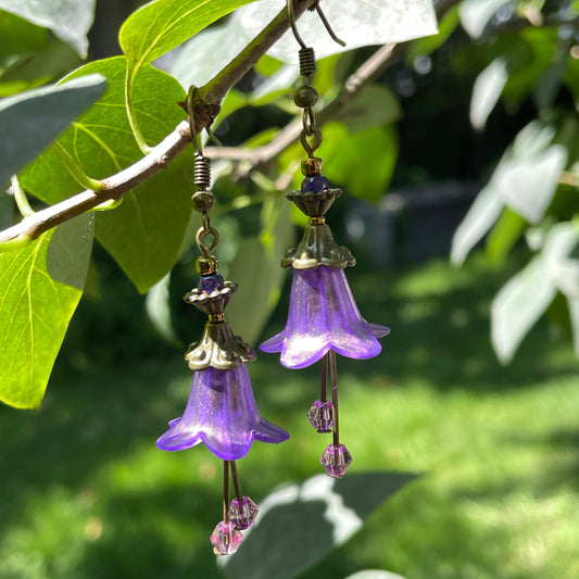 purple bell flower earrings with antique  bronze and purple beads. Purple crystal stamens. Earrings are hanging on a branch of a tree.