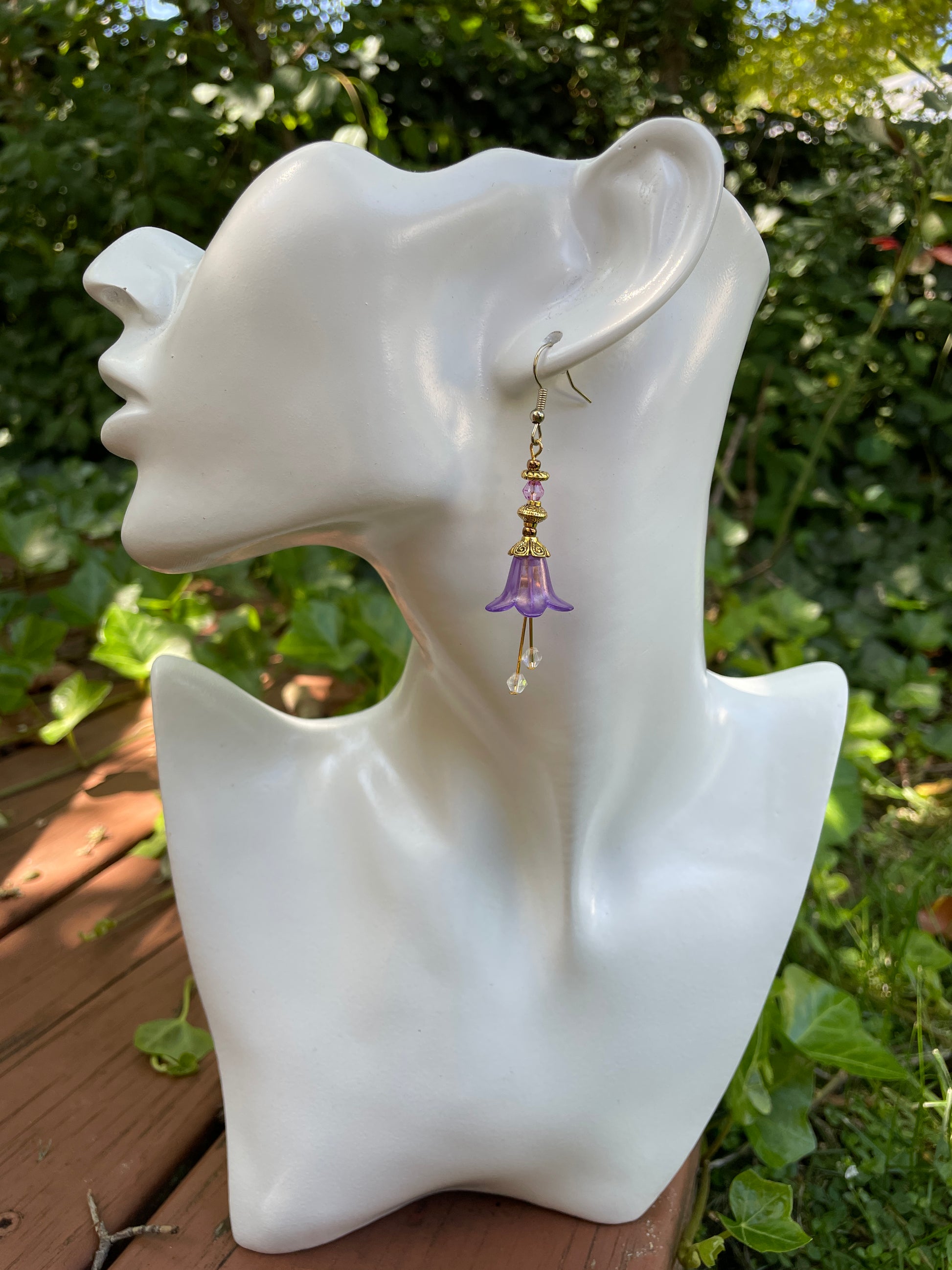 purple bell flower dangle earrings with clear crystal stamen and gold accent beads on a white model bust in a garden setting.