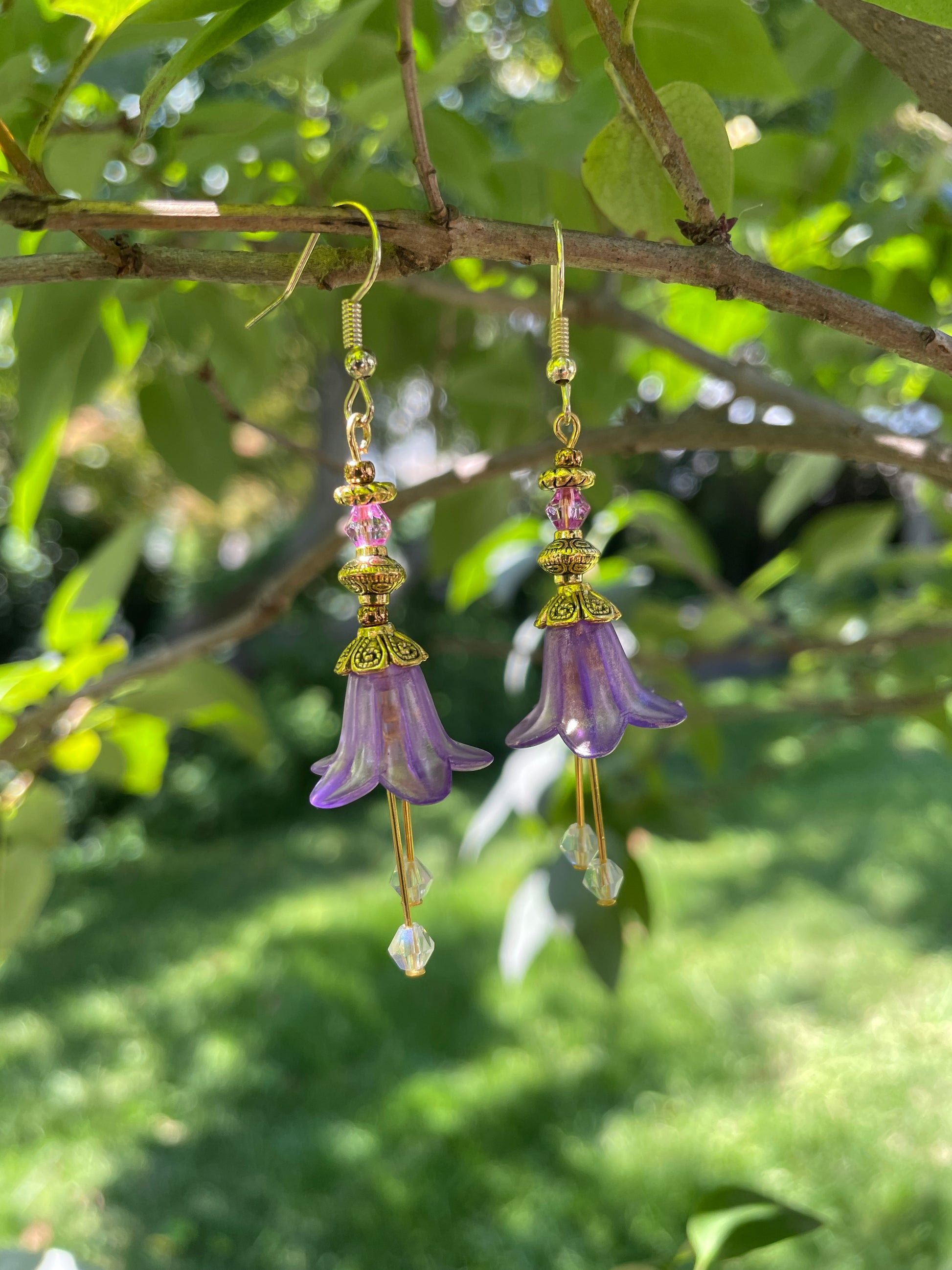 purple bell flower dangle earrings with clear crystal stamen and gold accent beads on a branch of a tree in a garden.