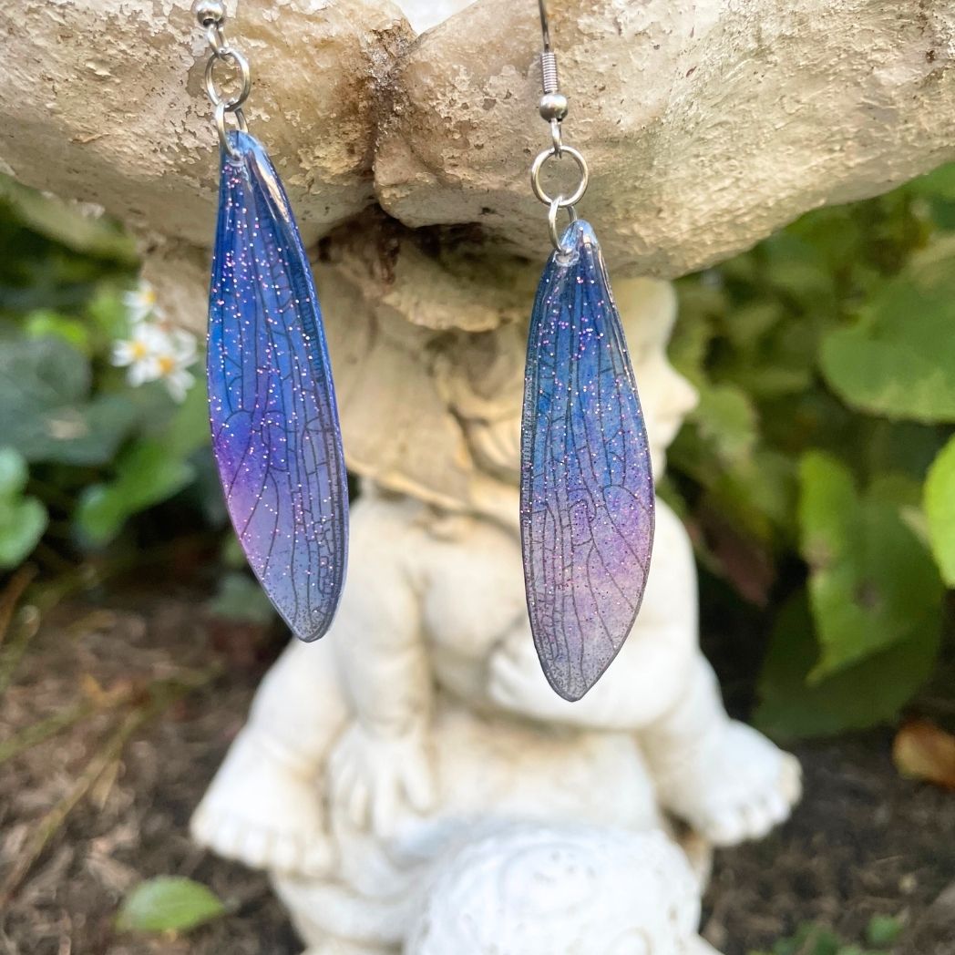 Sparkly glitter blue and purple dragonfly wing earrings hanging from a garden ornament.