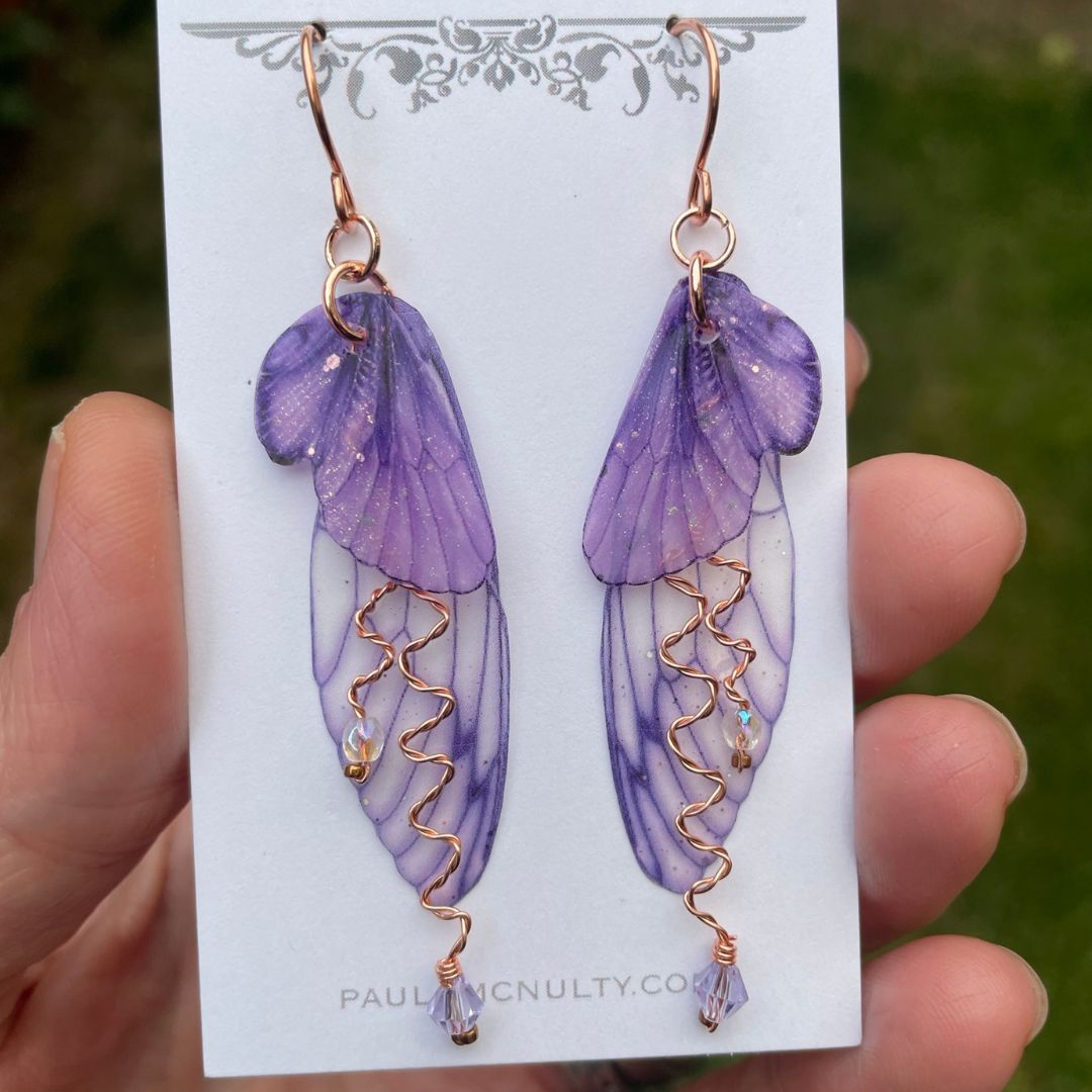 Violet purple fairy wing earrings with lavender crystals and copper wires. Displayed on a earring card held in a hand.