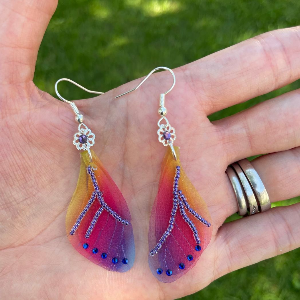 Colorful rainbow sheer fabric with beading and blue crystals. A flower bead with a crystal connects the wings to the ear wires. Display in a hand.