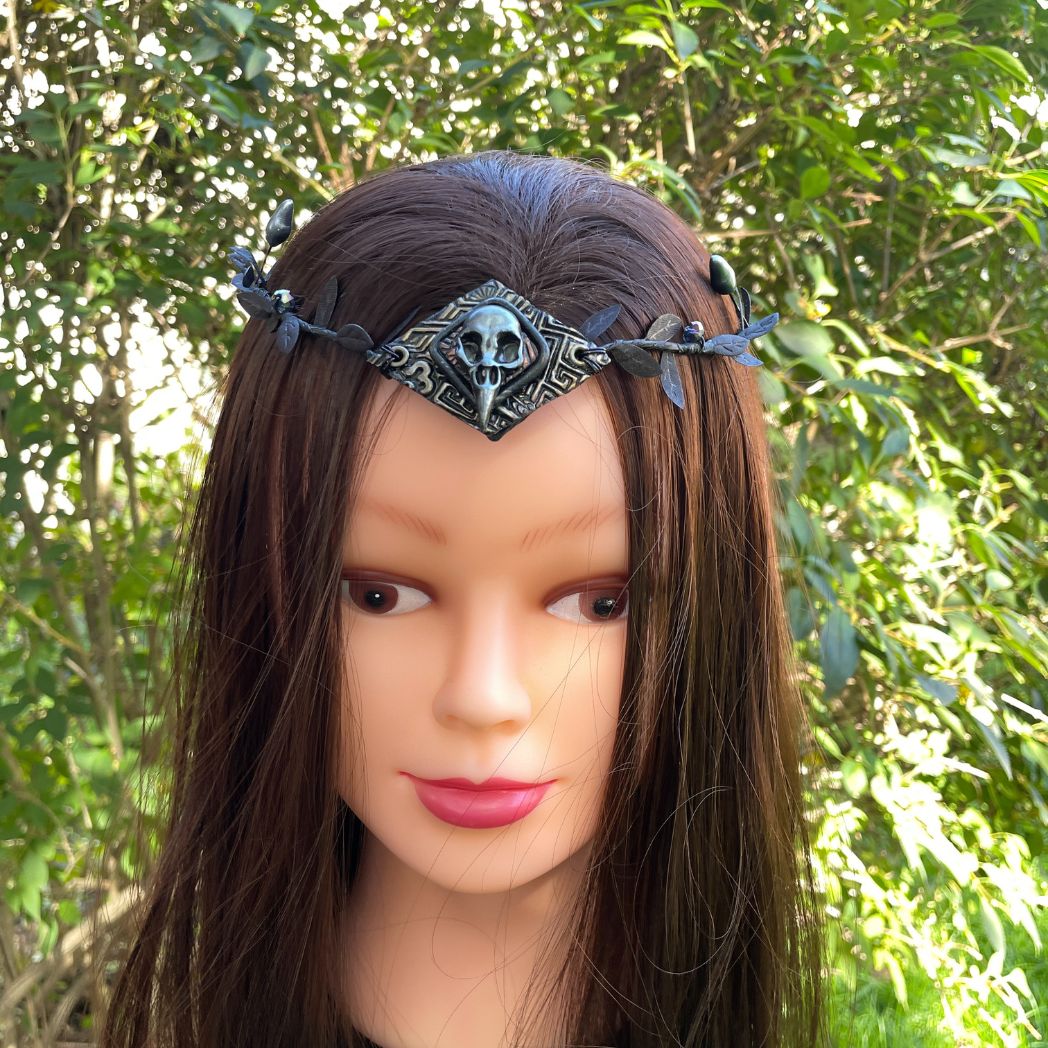 A silver raven skull on textured gold highlighted diamond shape focal on a black leaf circlet with black crystals and silver flower buds. Displayed on a model.