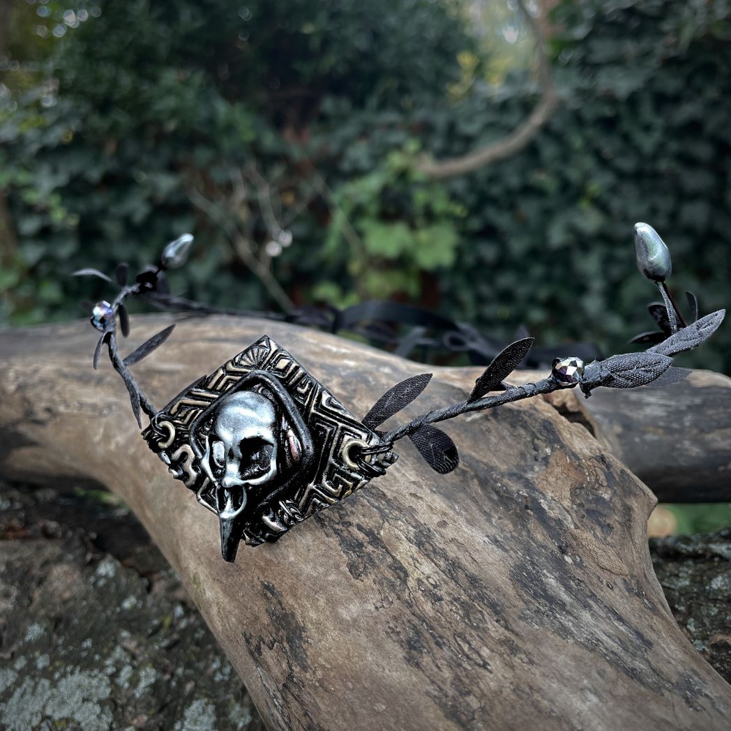 A silver raven skull on textured gold highlighted diamond shape focal on a black leaf circlet with black crystals and silver flower buds. Circlet is resting on a tree branch.