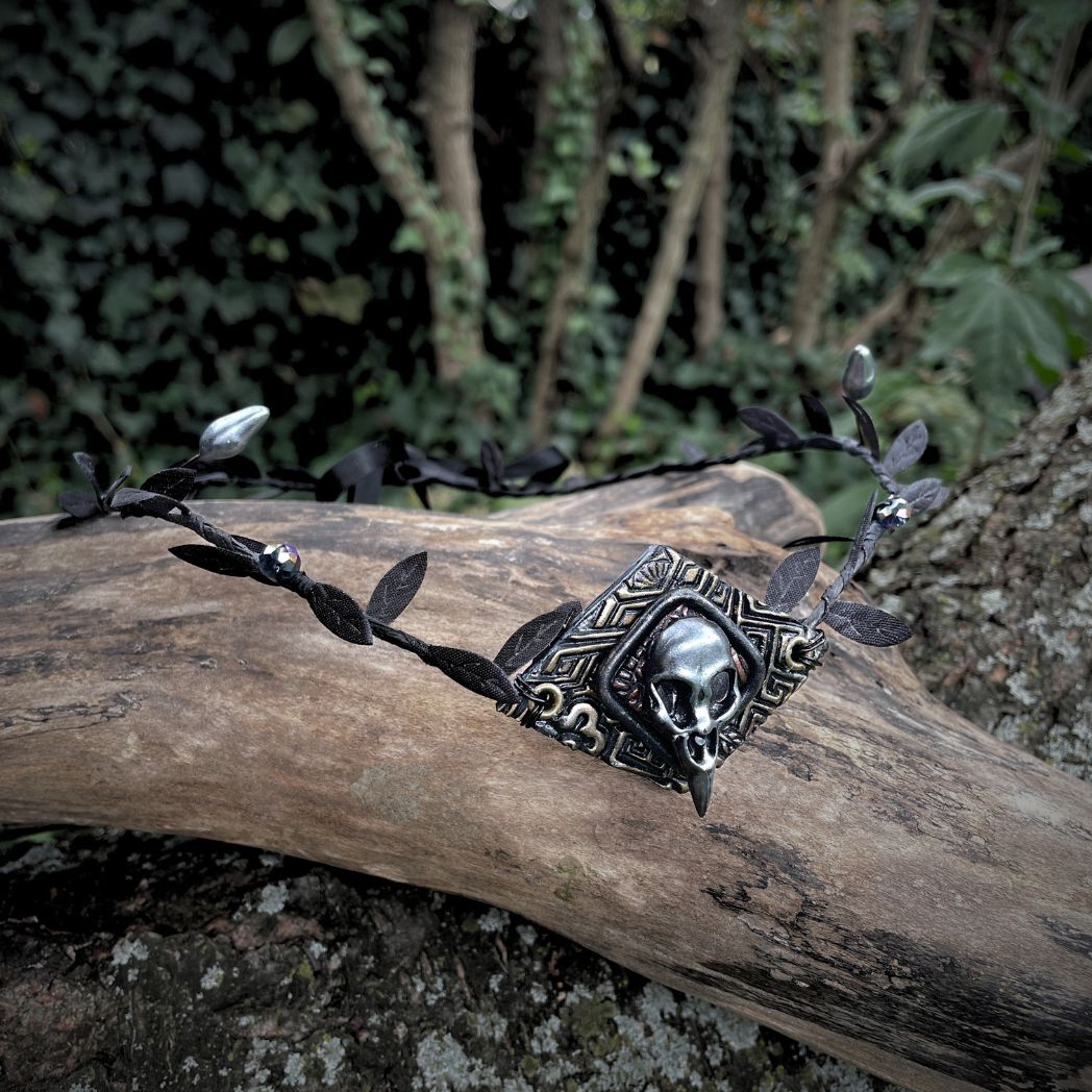 A silver raven skull on textured gold highlighted diamond shape focal on a black leaf circlet with black crystals and silver flower buds. Circlet is resting on a tree branch.