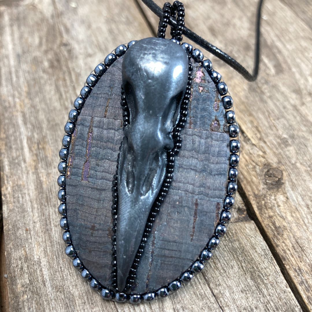 An oval pendant with a polymer clay crow skull on gray cork and hand beaded with silver and black seed beads on a wood background