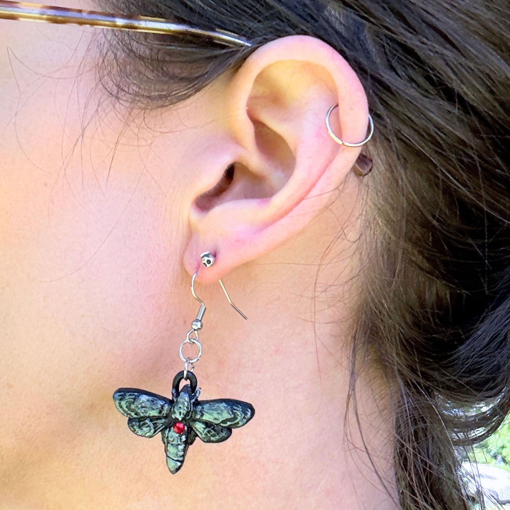 A close-up of a person's ear wearing a dangling moth charm earring. The earring has a silver-toned fishhook back, a silver ring connector, and a moth charm with dark green and black textured wings and a small red jewel at the center.