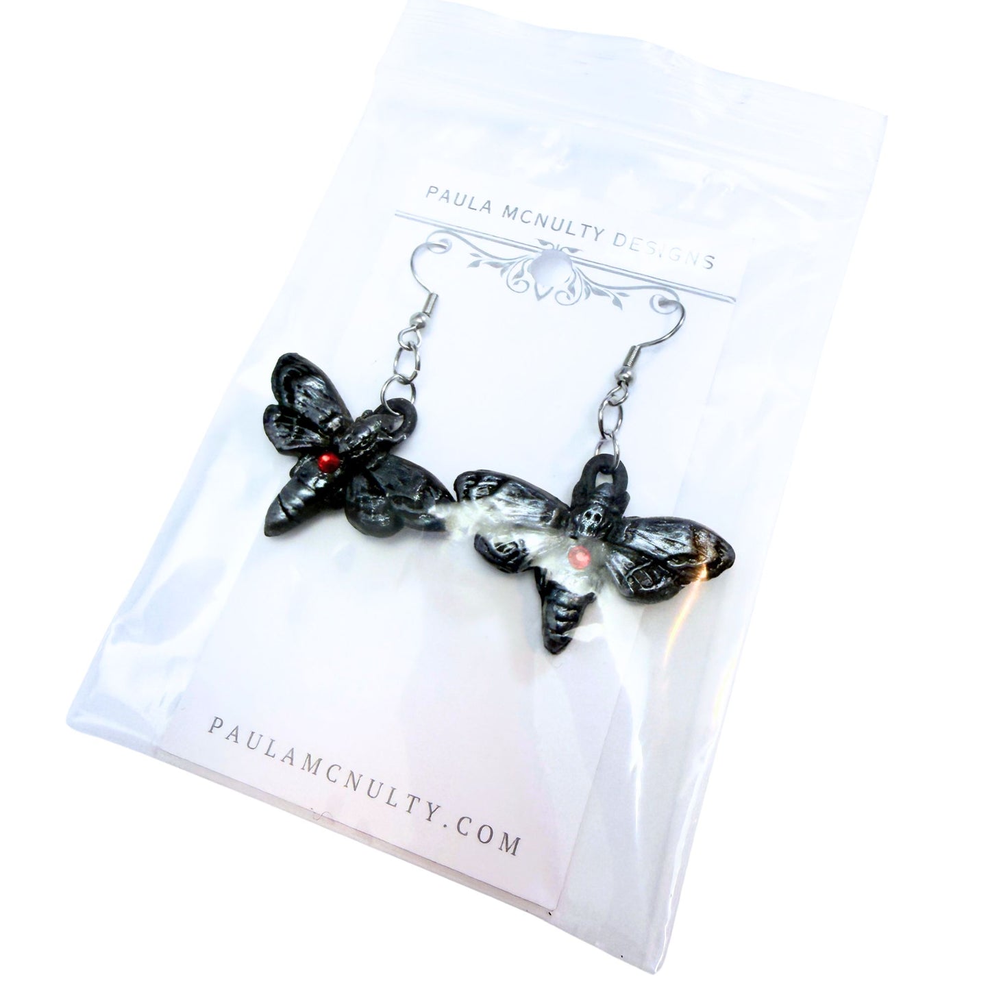 A pair of black gothic moth earrings with small red gemstone eyes, displayed on a white backing card printed with 'PAULA MCNULTY DESIGNS' and 'PAULAMCNULTY.COM'.