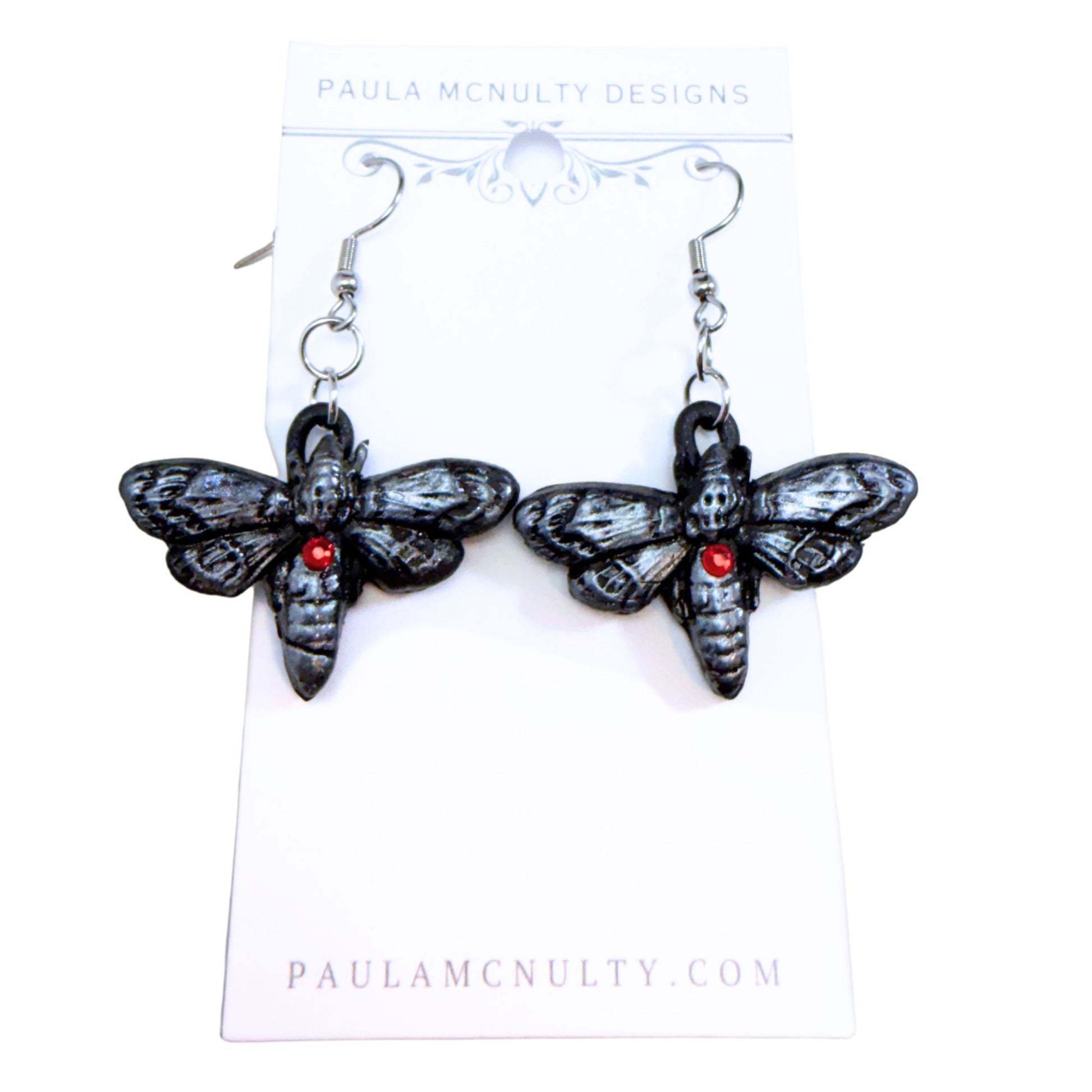 A pair of gothic-style black moth earrings with small red gemstone accents, hanging from silver fishhook wires on a white display card from Paula McNulty Designs.