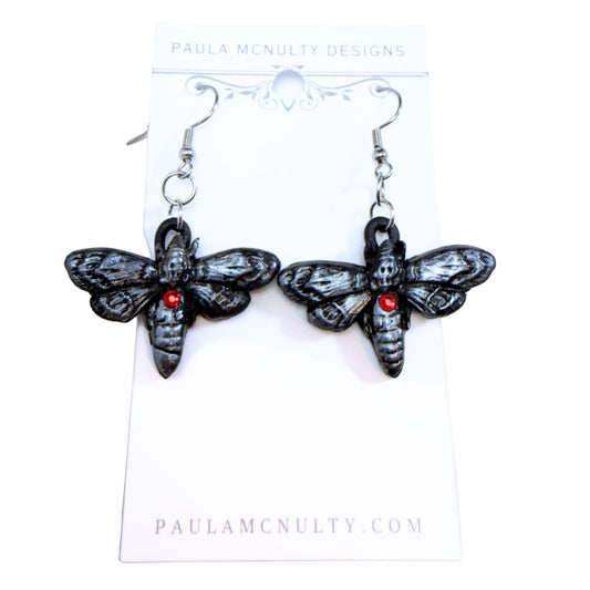 A pair of gothic-style black moth earrings with small red gemstone accents, hanging from silver fishhook wires on a white display card from Paula McNulty Designs.