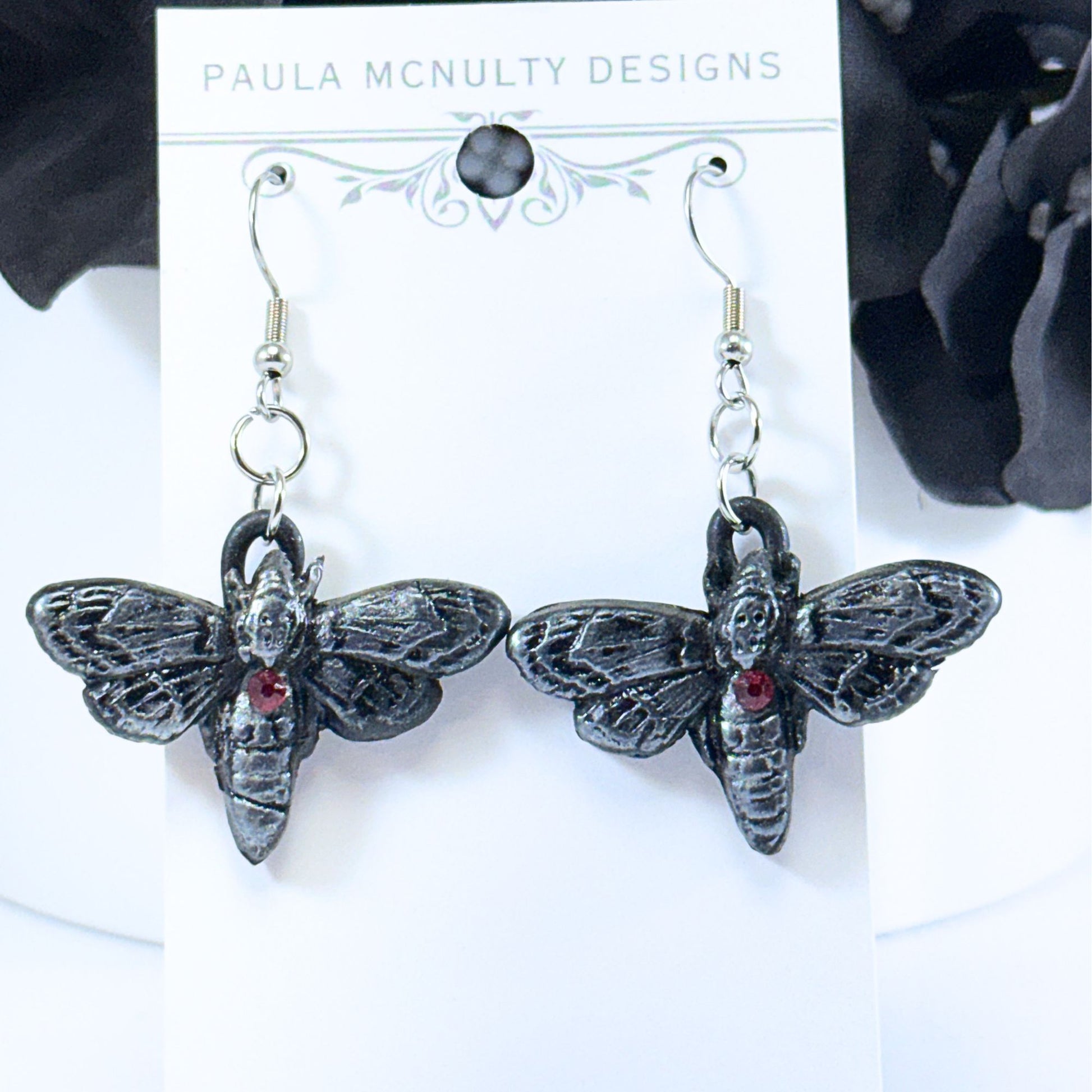 A pair of gothic-style bumblebee earrings by Paula McNulty Designs, featuring textured black metal wings and bodies with a small red gemstone on the abdomen, displayed on a white card.