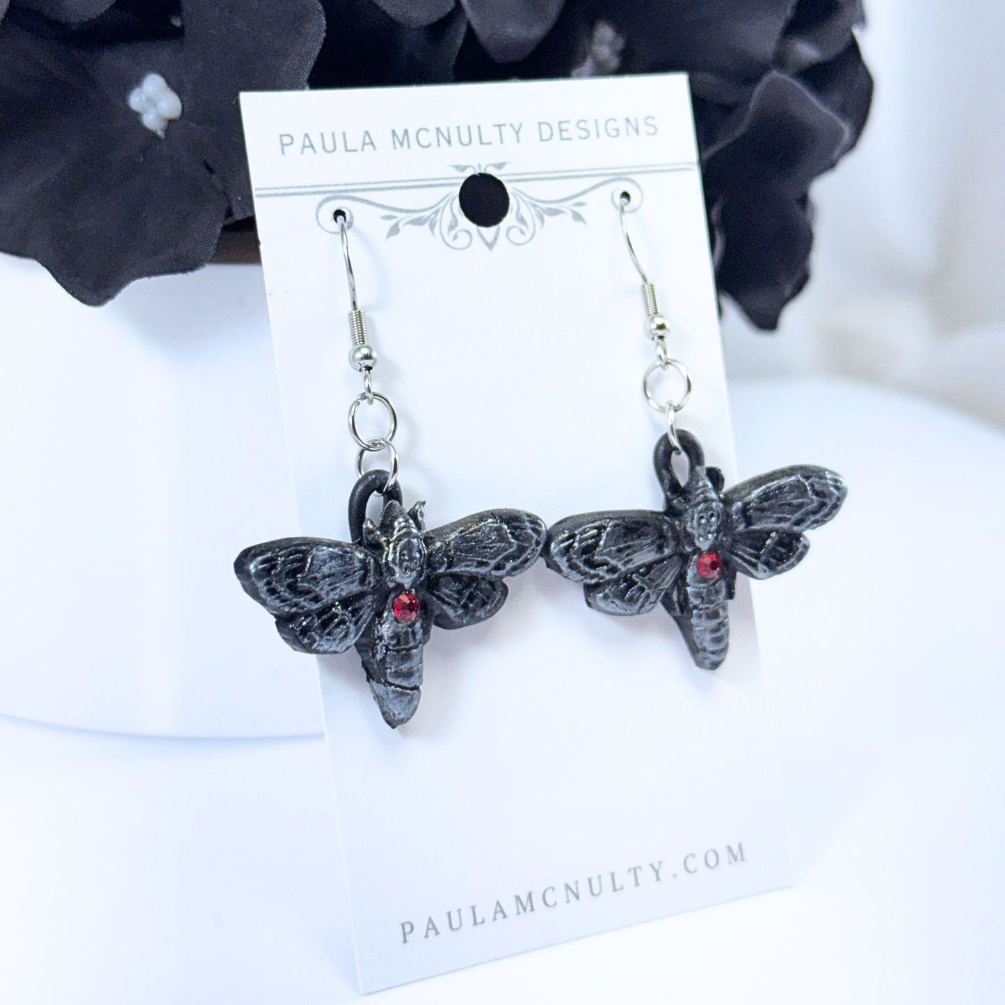 A pair of gothic moth earrings by Paula McNulty Designs, featuring textured black metal moths with small red gemstone eyes, displayed on a white card.